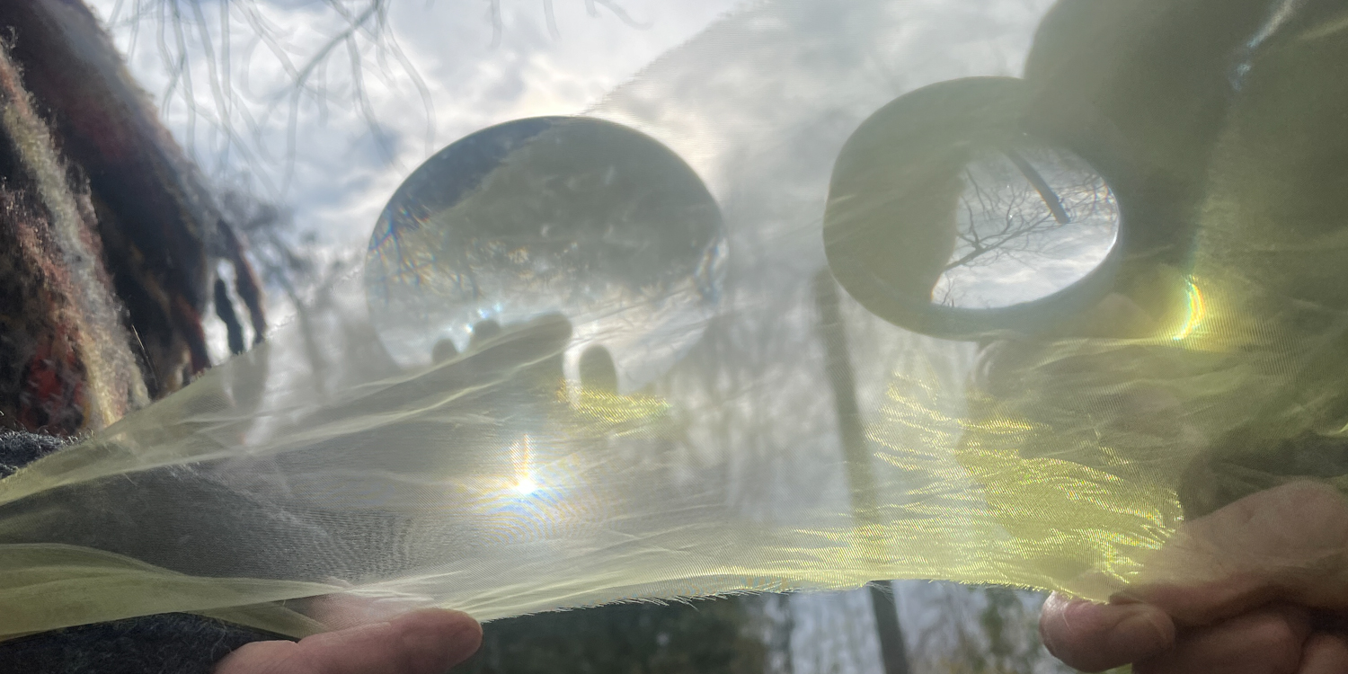 Close-up of hands holding translucent yellow fabric stretched under two glass lenses, with sunlight refracting through and creating small rainbow flares, against a backdrop of tree branches and cloudy sky.