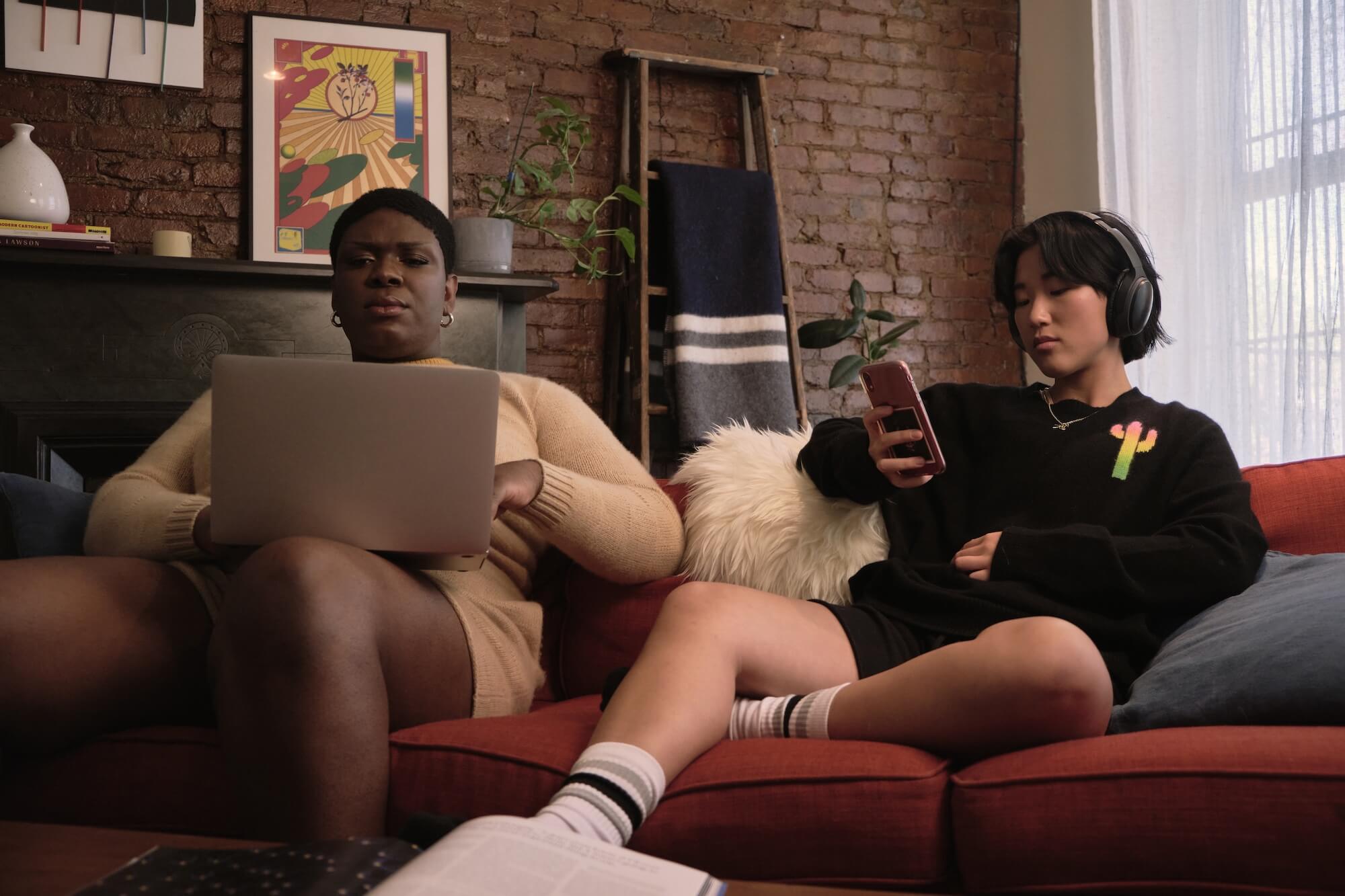 Two POC roommates on a red sofa. One is using a laptop, and the other is wearing headphones and looking at a phone.