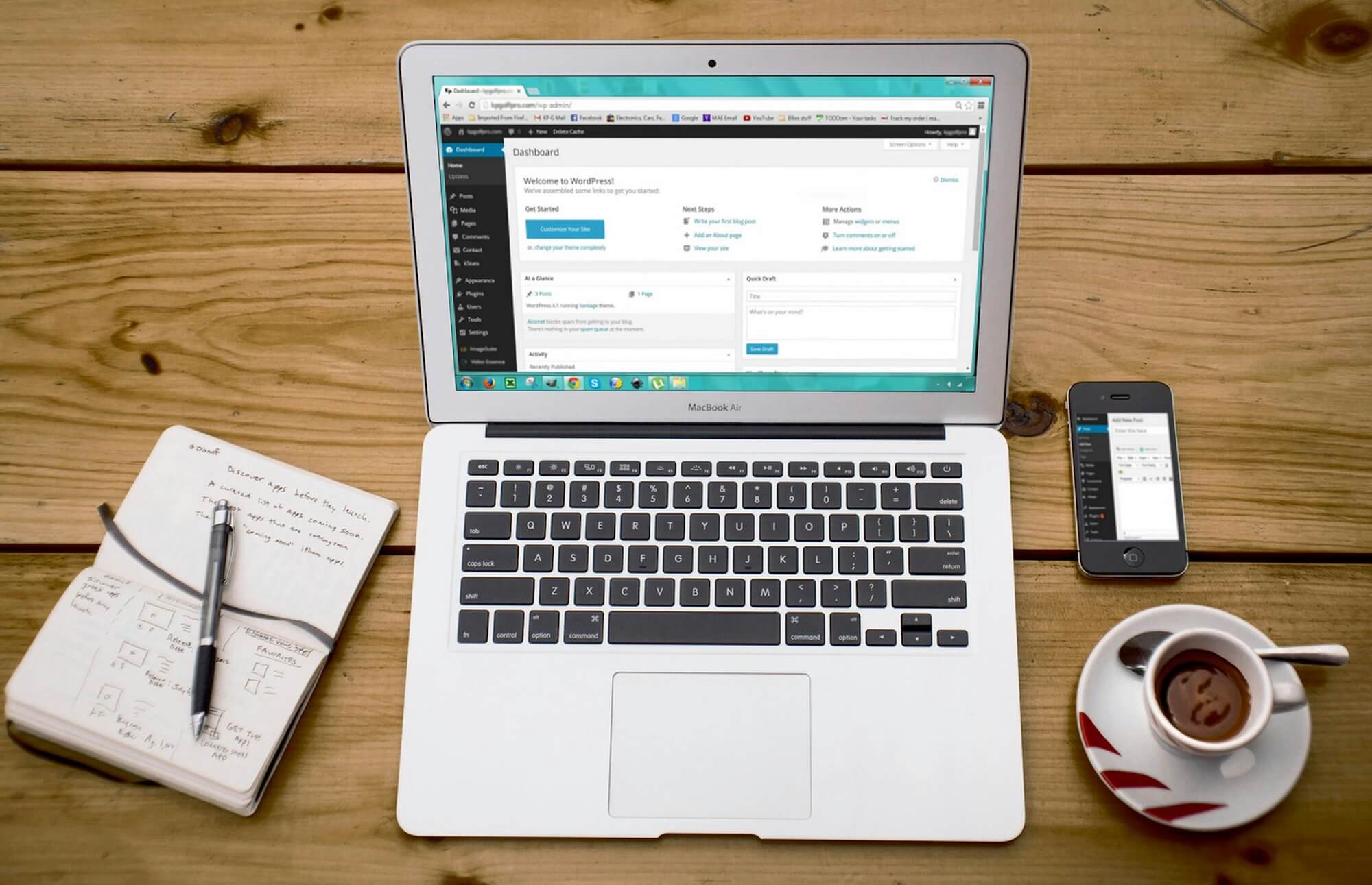 A turned-on MacBook Air with WordPress CMS on screen, sitting on a wooden desk beside a black iPhone 4, a cup of tea, and a notebook.