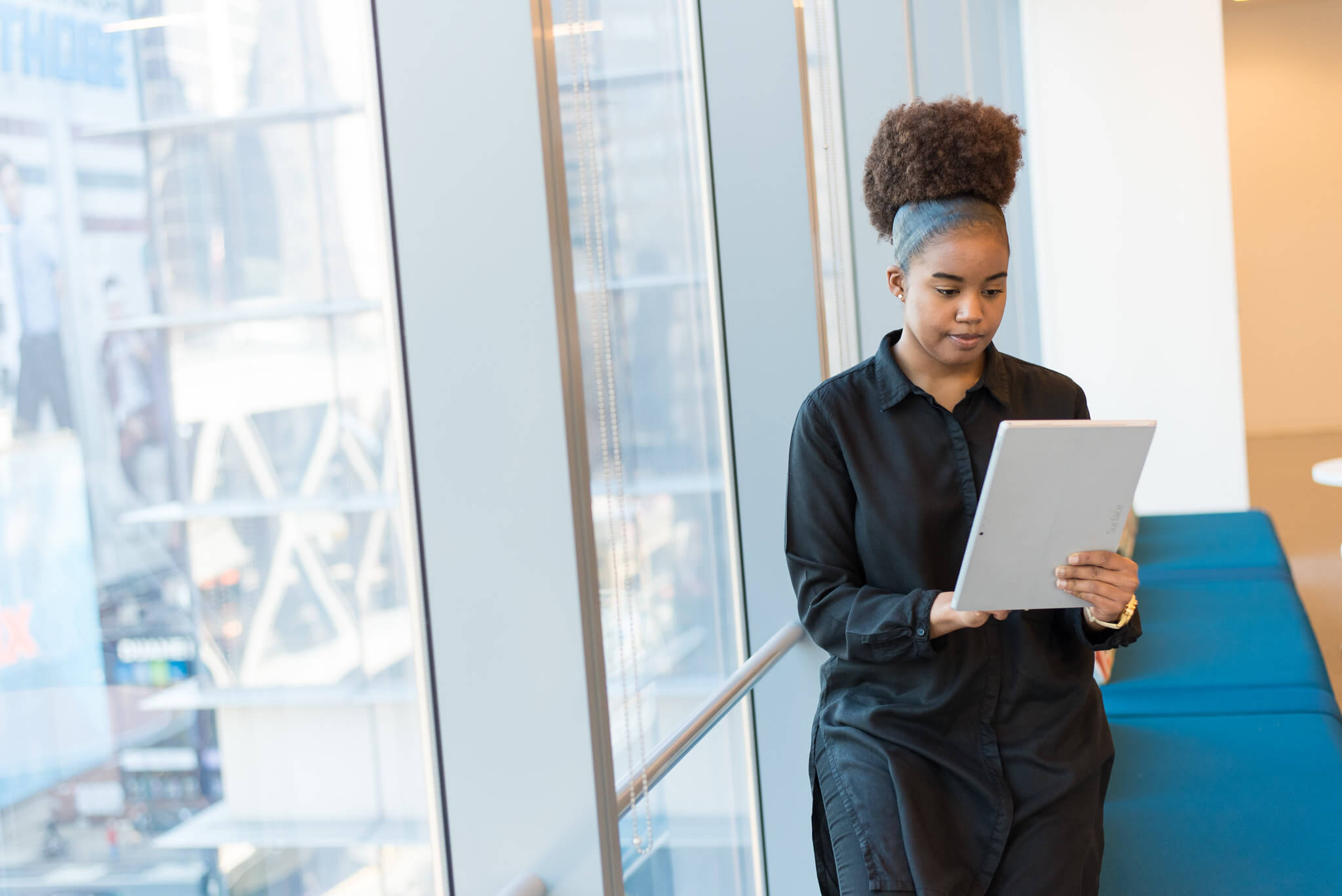 A person of colour wearing black standing next to full-length windows looking at the large tablet in their hand.