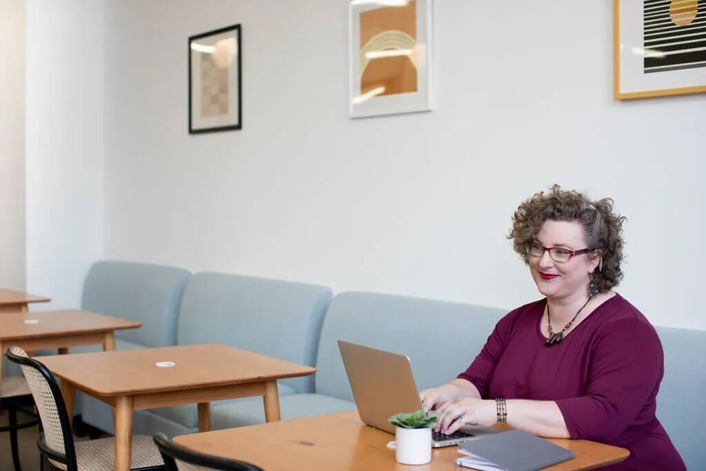 A white plus-size woman with curly hair working on a laptop at a table in a cafe.