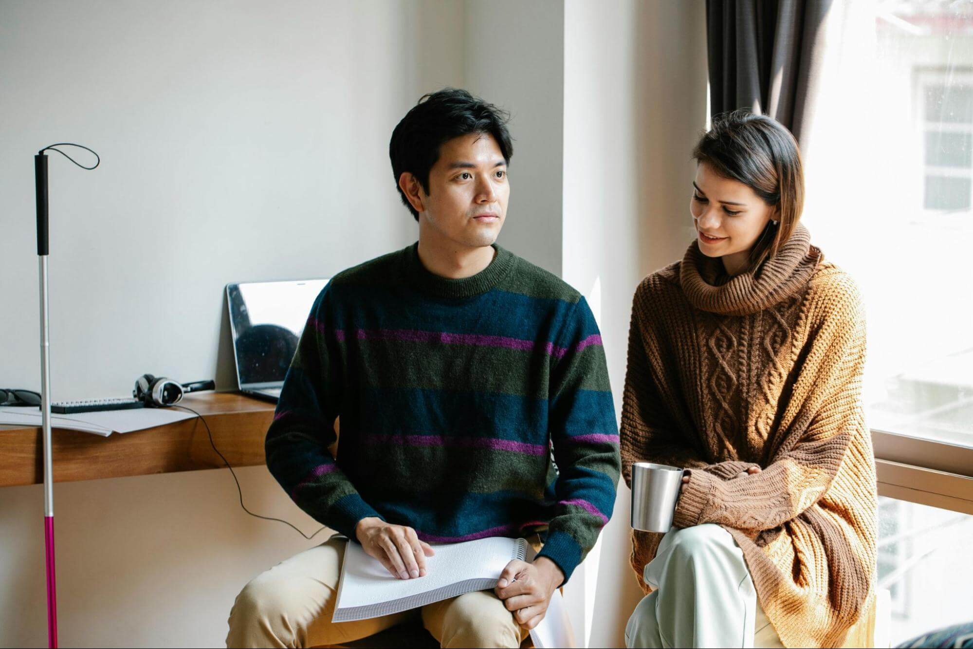 A blind man with dark hair wearing a blue, green, and pink striped jumper is reading Braille. A woman wearing a chunky knitted jumper is sitting beside him, drinking from a metallic mug.