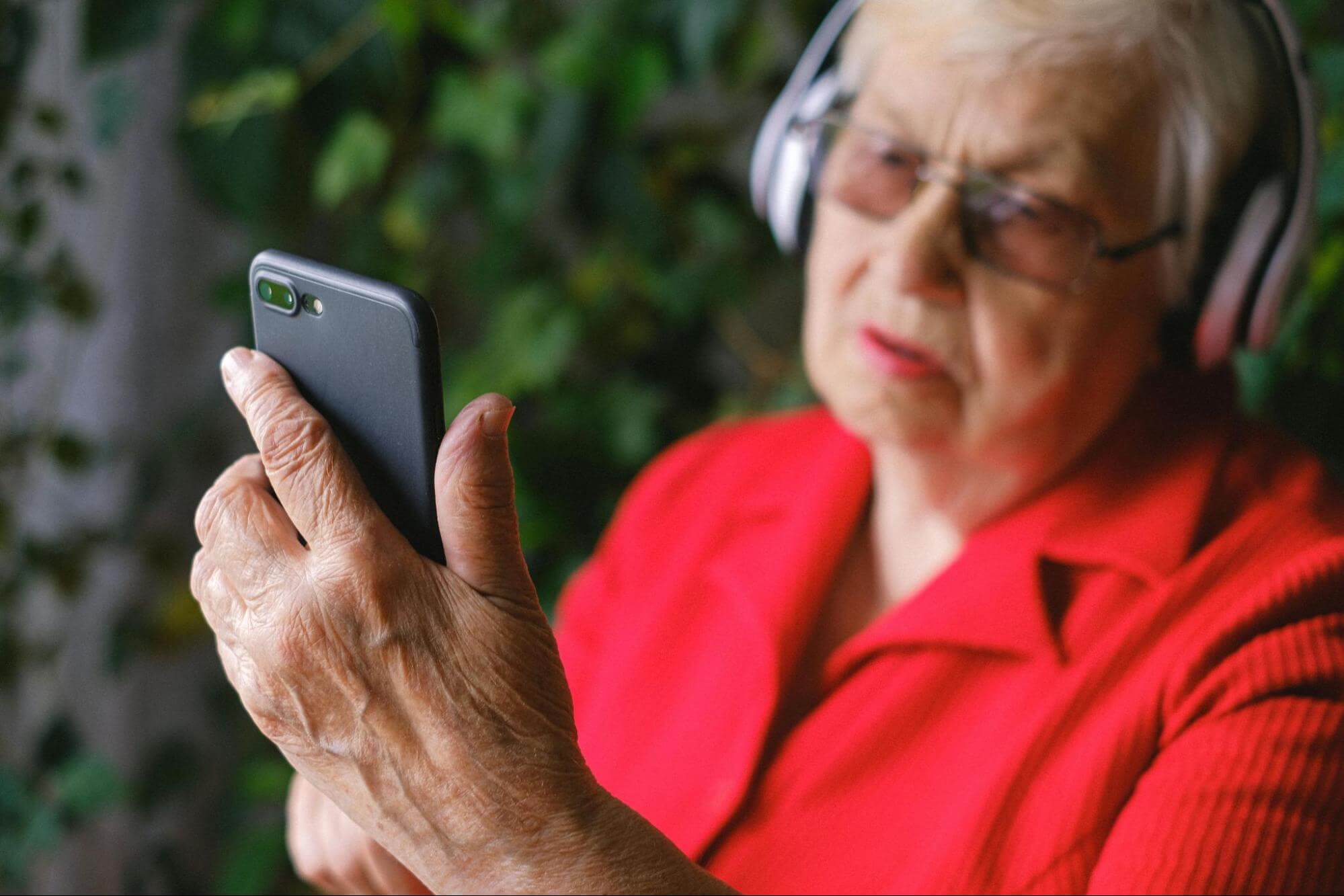 An older woman with grey hair wearing a red shirt, glasses, and silver over-ear headphones is holding a smartphone and looking at the screen.