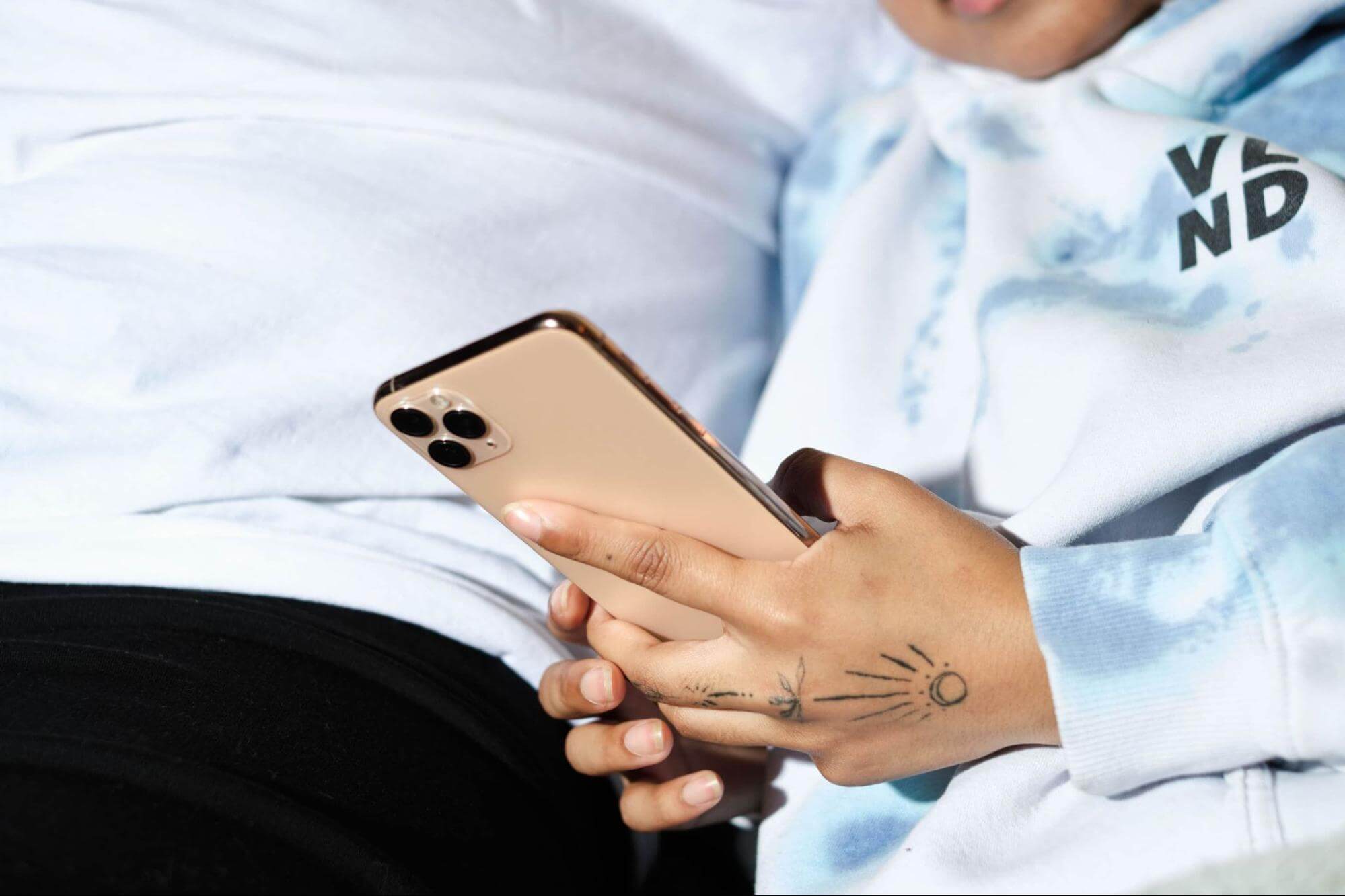 A tattooed hand holding a smartphone. The phone-holding person is wearing a blue tie-dyed hoodie and the person sitting next to them is wearing a white t-shirt. The image is cropped so we can’t see their faces, only their torsos.