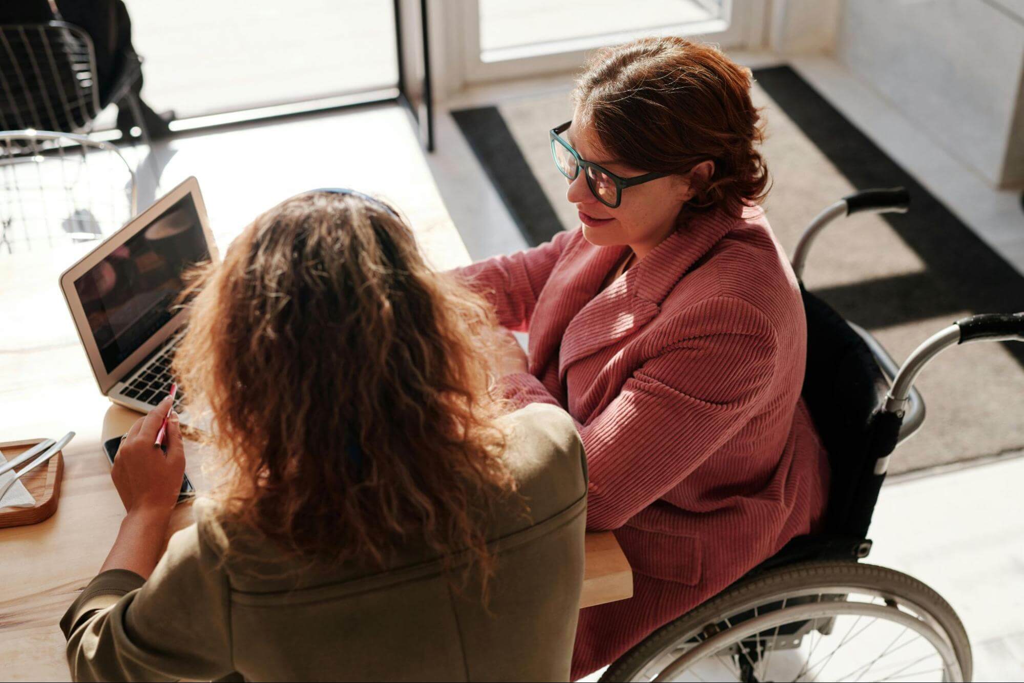 Two people are sitting at a desk using a laptop together. One is wearing a khaki jacket and has their back to the camera, and the other is dressed in pink and is sitting in a wheelchair, side-on to the camera