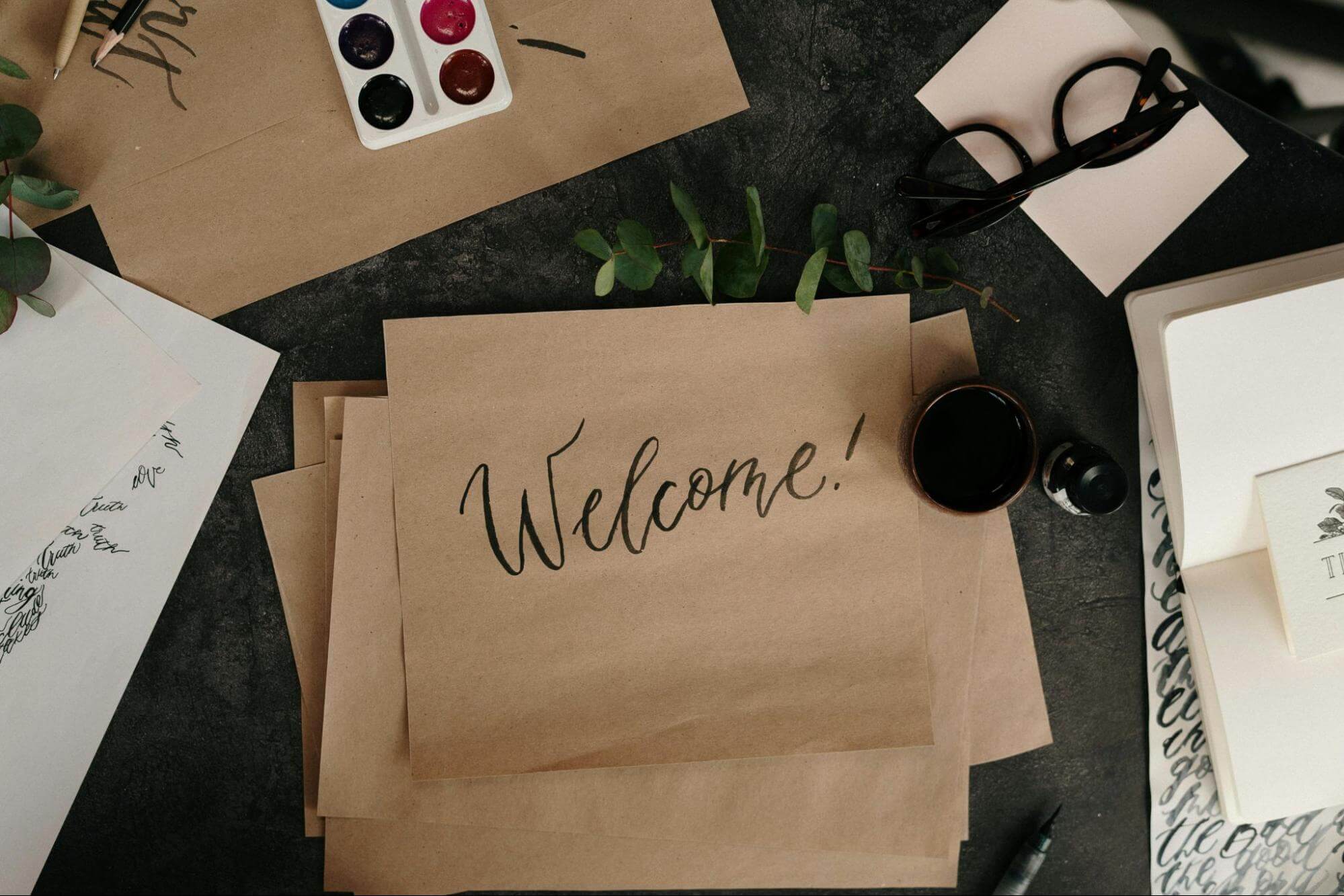 A desk covered in brown and white paper, a notebook, a pair of black glasses, and some paints. The piece of paper in the centre of the table is brown and has the word “welcome” handwritten on it.