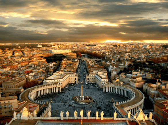 Aerial sunset panorama of St. Peter’s Square and the surrounding cityscape of Rome, with golden light illuminating the Vatican colonnades and distant rooftops.