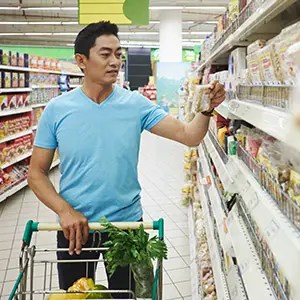 A man in a blue shirt is shopping in a grocery store. He is holding a package and examining it while pushing a cart filled with groceries, including leafy greens. Shelves stocked with various products are visible in the background.