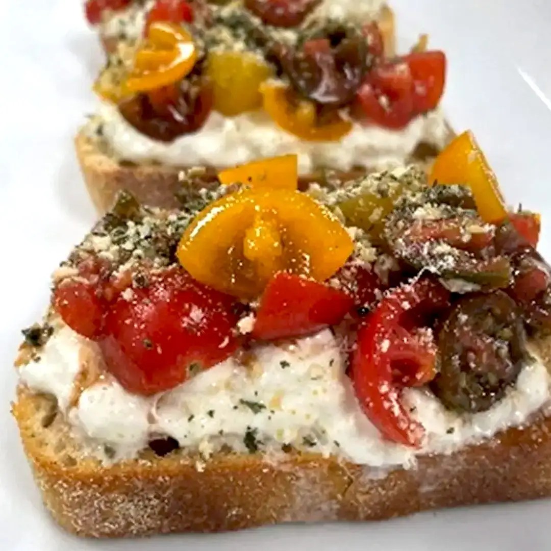 Three slices of toasted bread topped with creamy white cheese, chopped red and yellow tomatoes, herbs, Pesto Seasoning, and a sprinkle of grated cheese are arranged in a row on a white plate.