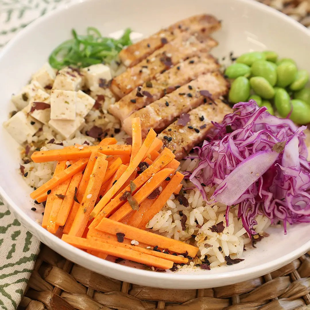 A bowl of rice topped with grilled chicken slices, diced tofu, shredded purple cabbage, edamame, and julienned carrots. The dish is garnished with sesame seeds and seasonings, set on a woven mat background.