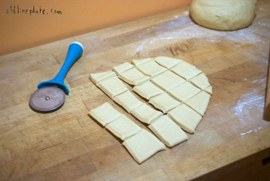 Cutting squares of fastnacht dough with pizza cutter on floured wooden board before deep frying