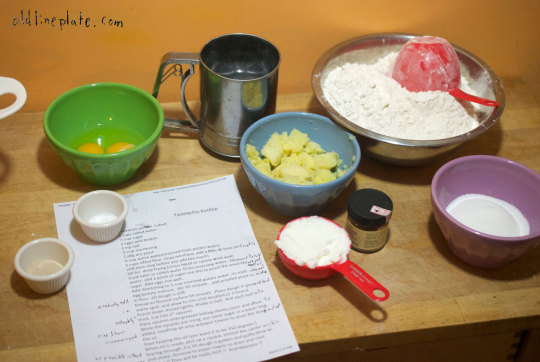 Baking ingredients for Fastnachts Küchlie including flour, sugar, yeast, eggs, shortening, and potato with recipe card on wooden counter