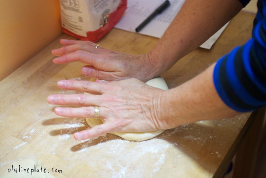 Two hands kneading dough on floured wooden surface, preparing Fastnachts Küchlie dough