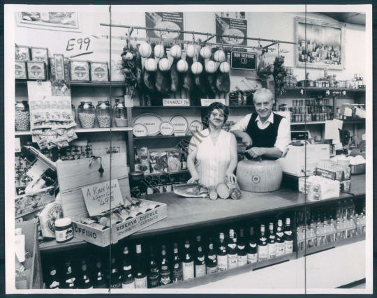 Vintage butcher shop display with two people behind counter, hanging meats, bottled goods, and products on shelves from Mastellone's Italian market