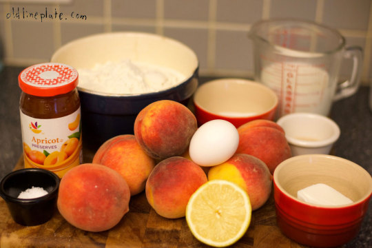 Ingredients for Baltimore Peach Cake laid out on counter including fresh peaches, apricot jam, eggs, flour, milk and lemon