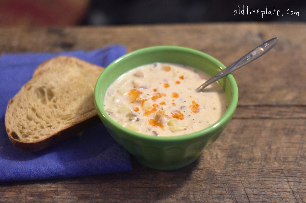 Creamy rockfish chowder in green bowl with bread slice, topped with bacon bits and herbs on wooden table