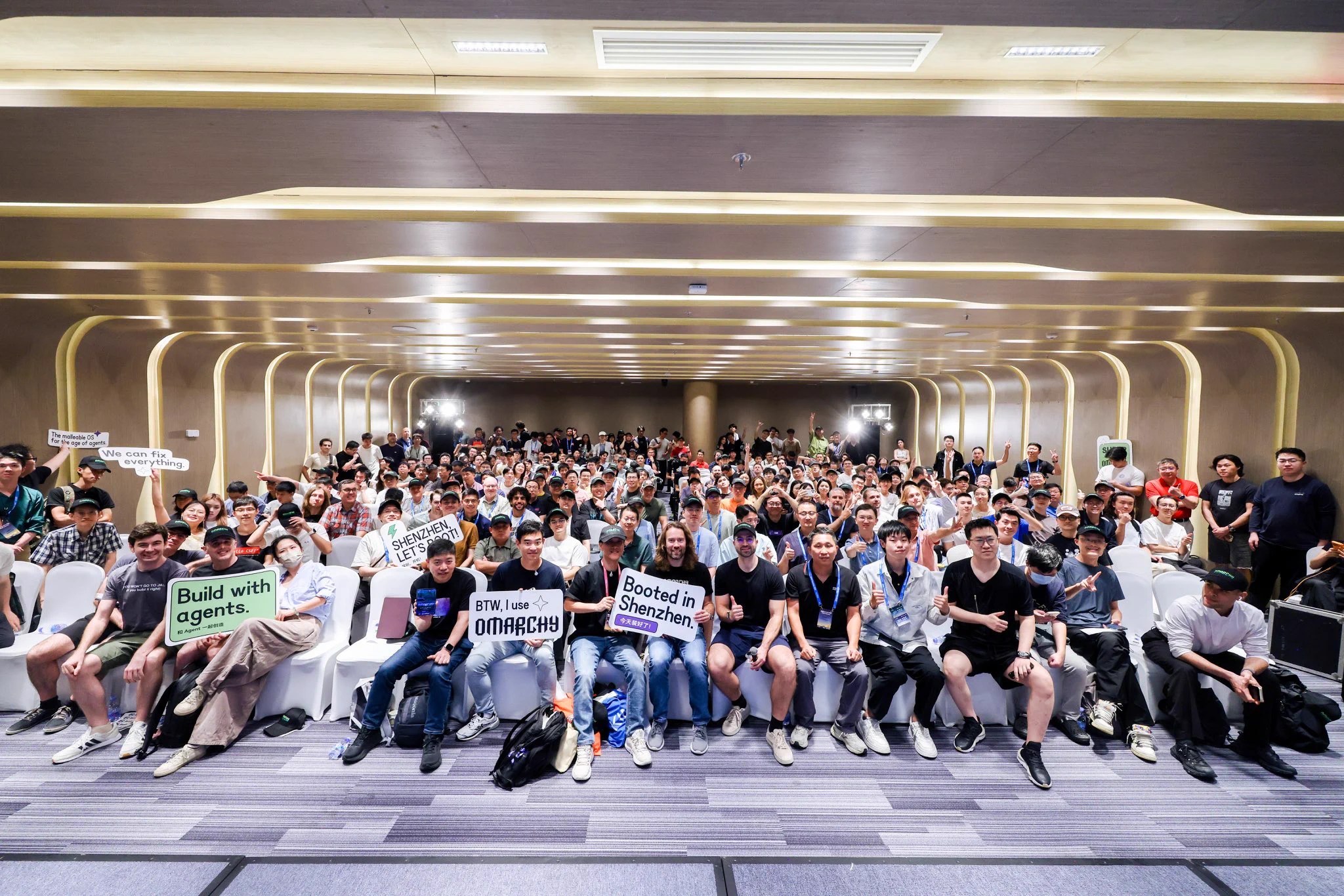 Group photo of a packed conference room at the Omarchy meetup in Shenzhen, with attendees in Omarchy caps holding signs reading Booted in Shenzhen, BTW I use Omarchy, Build with agents and Shenzhen, let's boot
