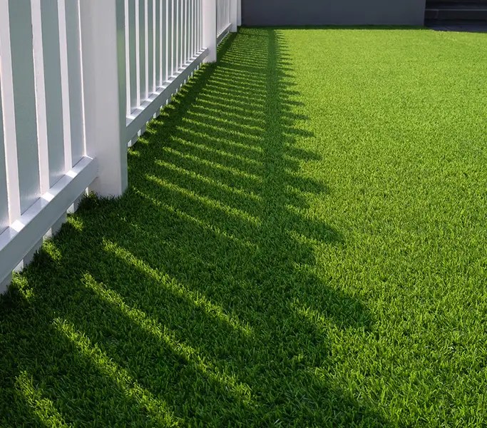 Sunlight and shadow of white wooden fence on green artificial turf surface in front yard of home, selective focus with high angle view