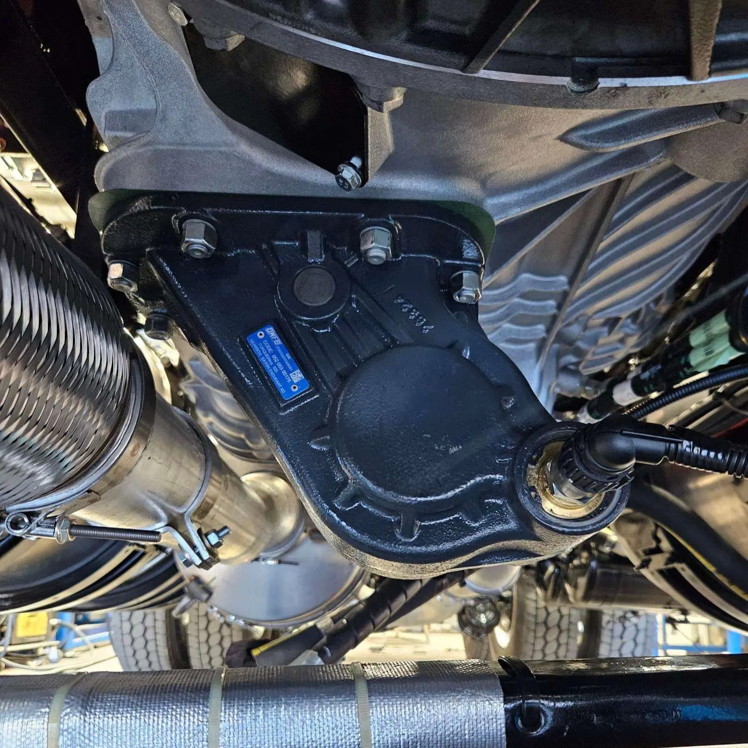 Installed OMFB Pacific ENDURANT XD PRO PTO shown mounted beneath a truck, featuring a durable black housing and visible sensor connection. Designed for seamless integration with transmission systems in heavy-duty vehicles.