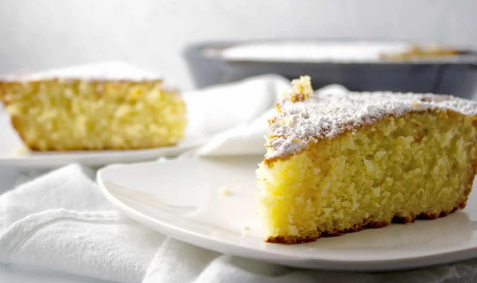A slice of yellow sponge cake dusted with powdered sugar on a white plate, with a whole cake and another slice in the background.