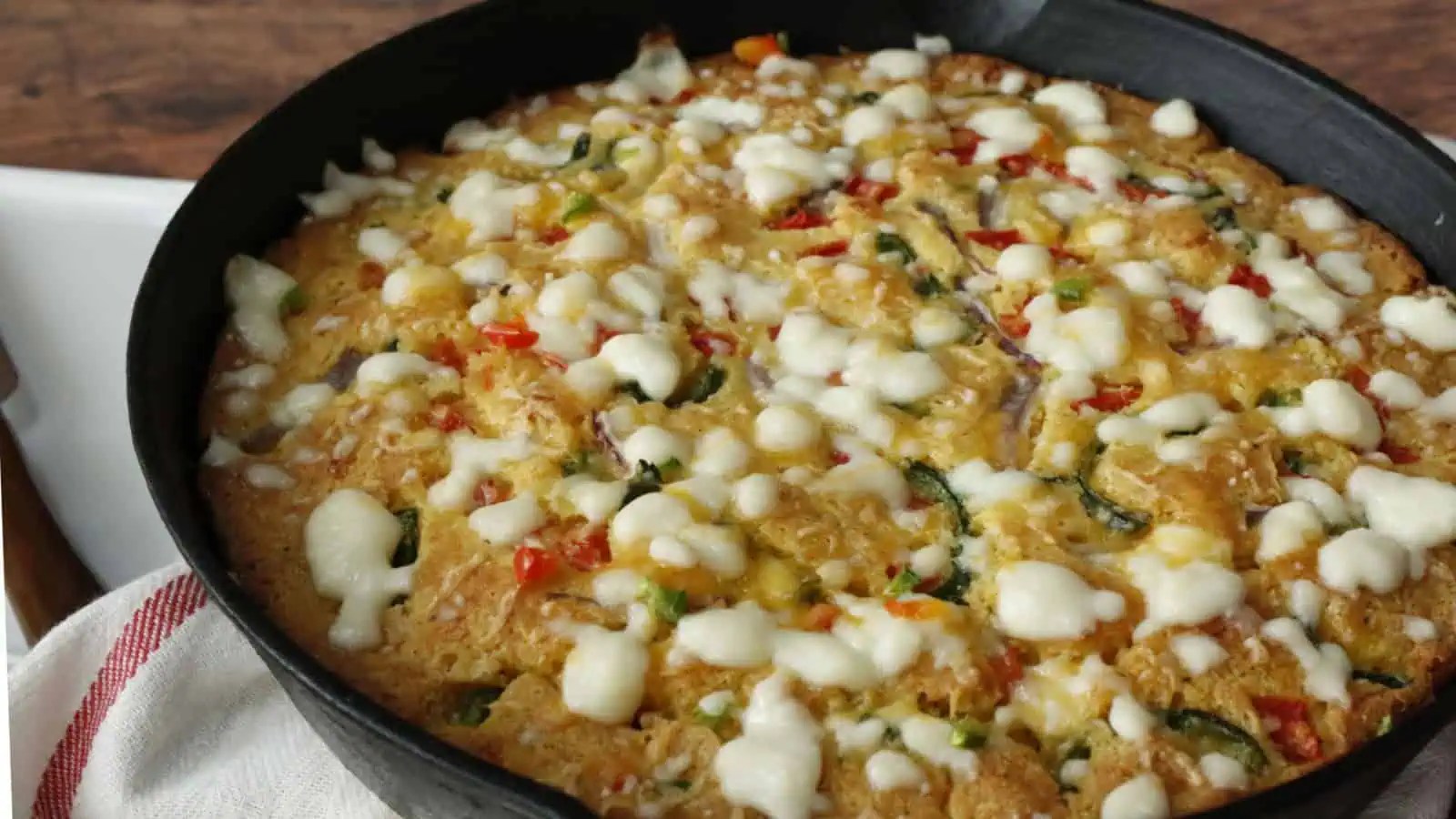 A baked cornbread dish in a black skillet, topped with melted cheese and visible pieces of red and green vegetables.