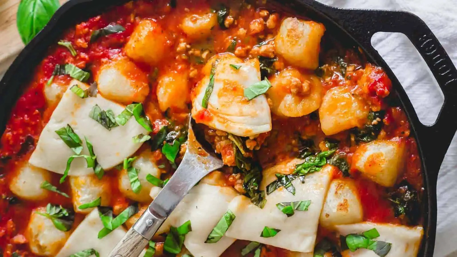 A skillet of cooked gnocchi in tomato sauce topped with chopped basil leaves and pieces of white rolled cheese. A metal spatula is lifting a serving from the skillet.