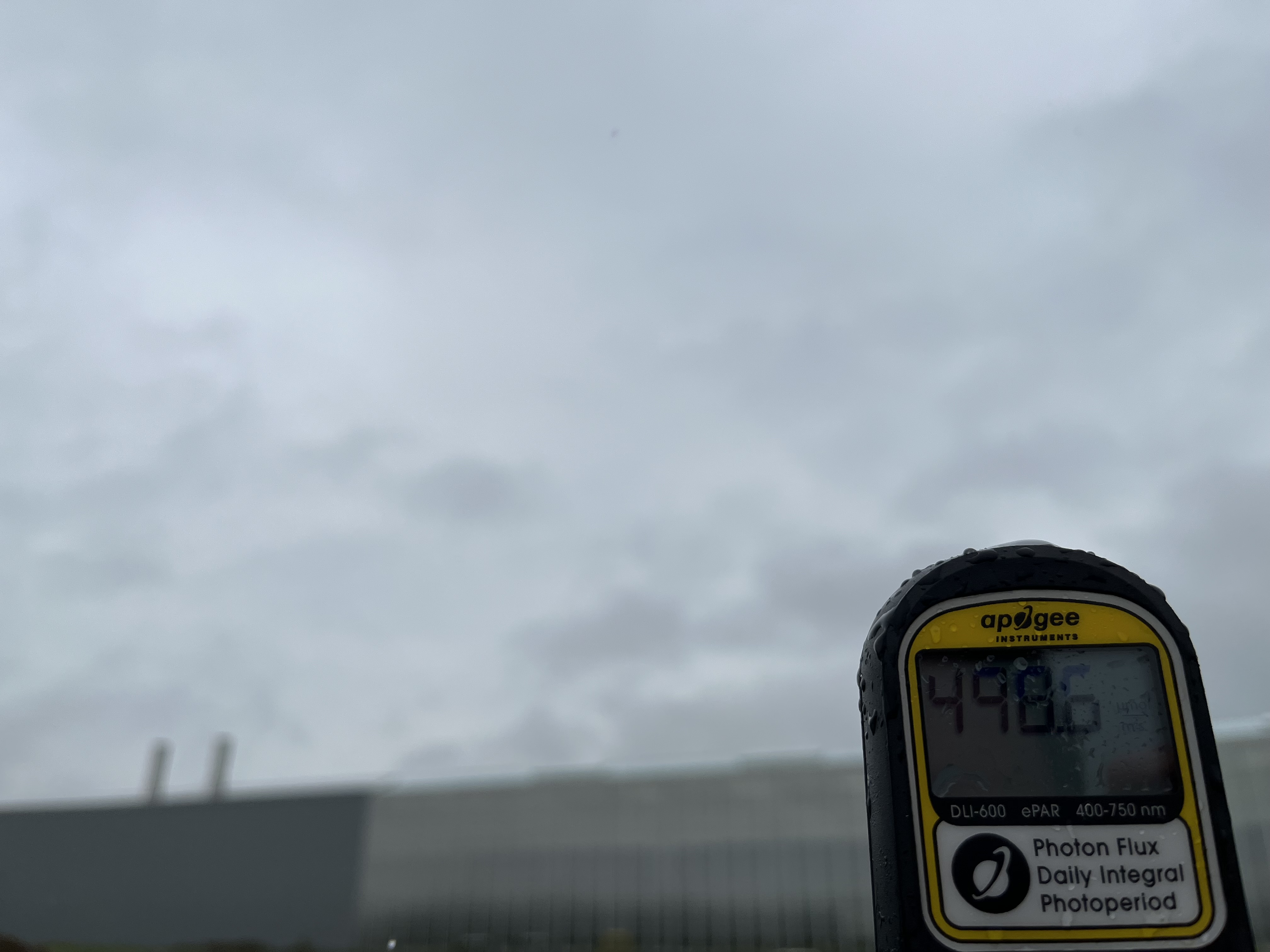 DLI meter in the forefront with a greenhouse under cloudy sky in the background