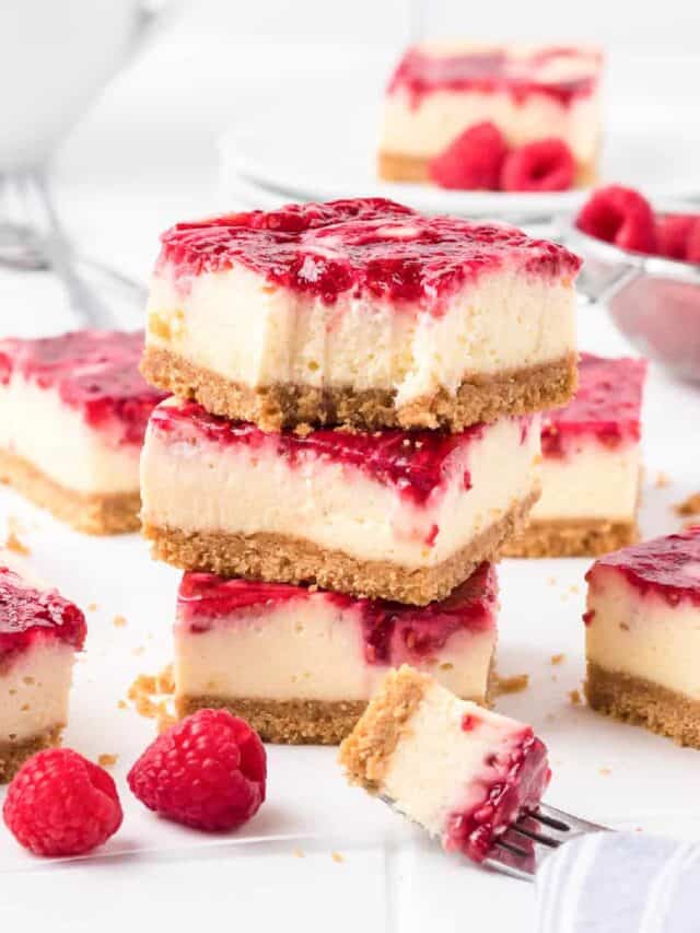 Stacked raspberry swirl cheesecake bars with the top bar missing a bite. Fresh raspberries and more cheesecake bars sit behind and nearby on the counter.