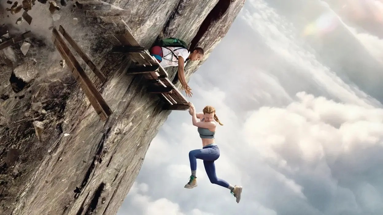 Harriet Slater as Jax hangs below a splintered wooden plank walkway bolted into a sheer cliff face, reaching up toward Arsema Thomas as Luce, who leans out from a rock crevice wearing a green backpack, with thick cloud cover filling the drop beneath them, in Fall 2: Deadpoint (2026).