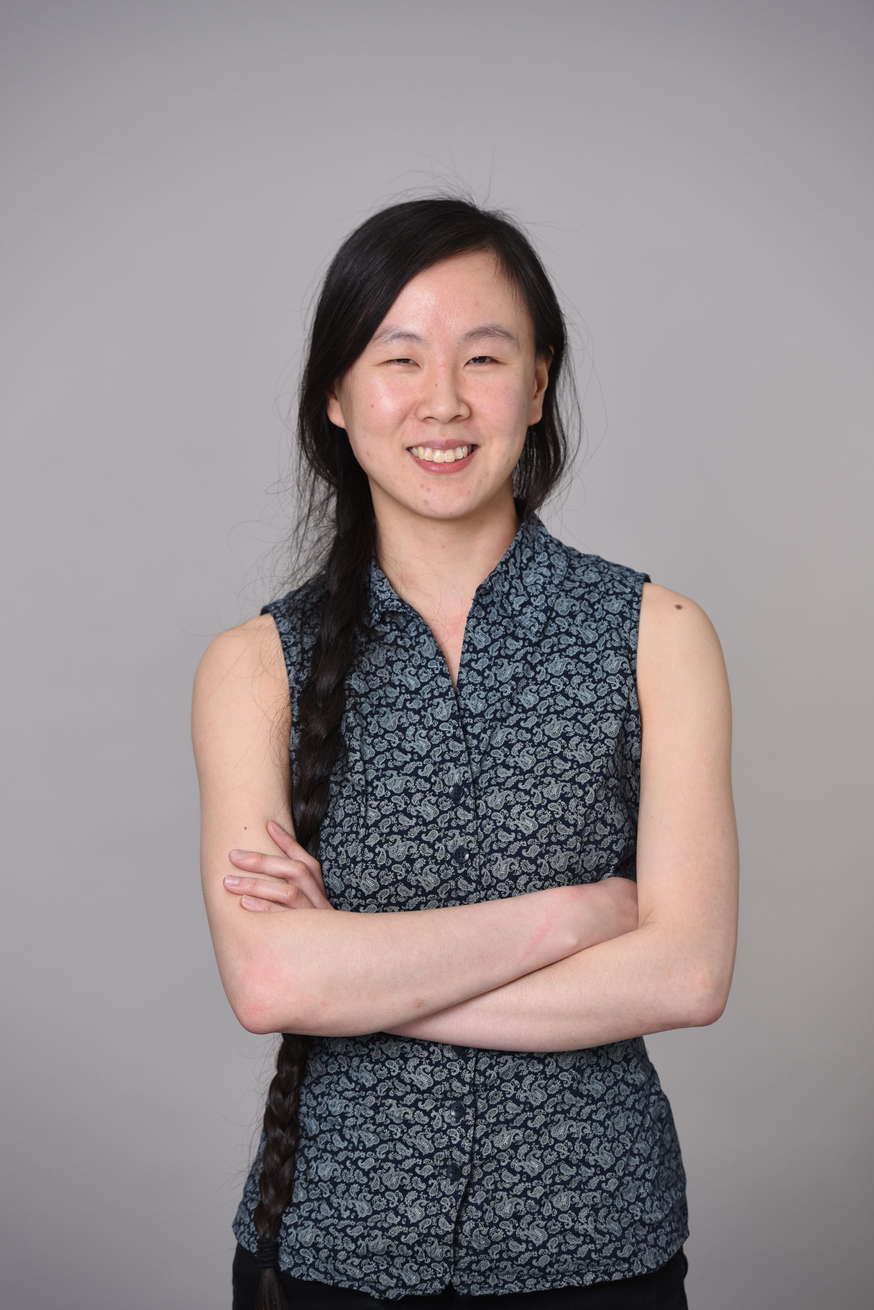A photo of Yan Kay Ho, standing in a blue-grey tunic with her arms crossed in front of her chest.