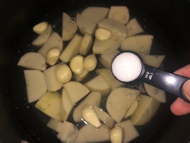 Adding salt to a pot with chopped potatoes and garlic cloves in it.
