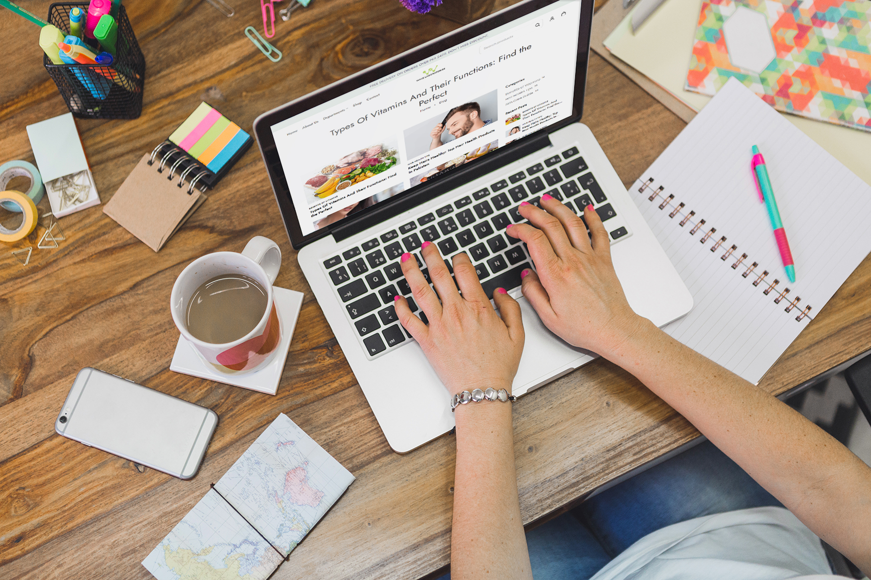 Overhead view of hands typing on a laptop displaying a webpage about vitamins, with a mug of coffee, smartphone, notebook, and stationery on a wooden desk.