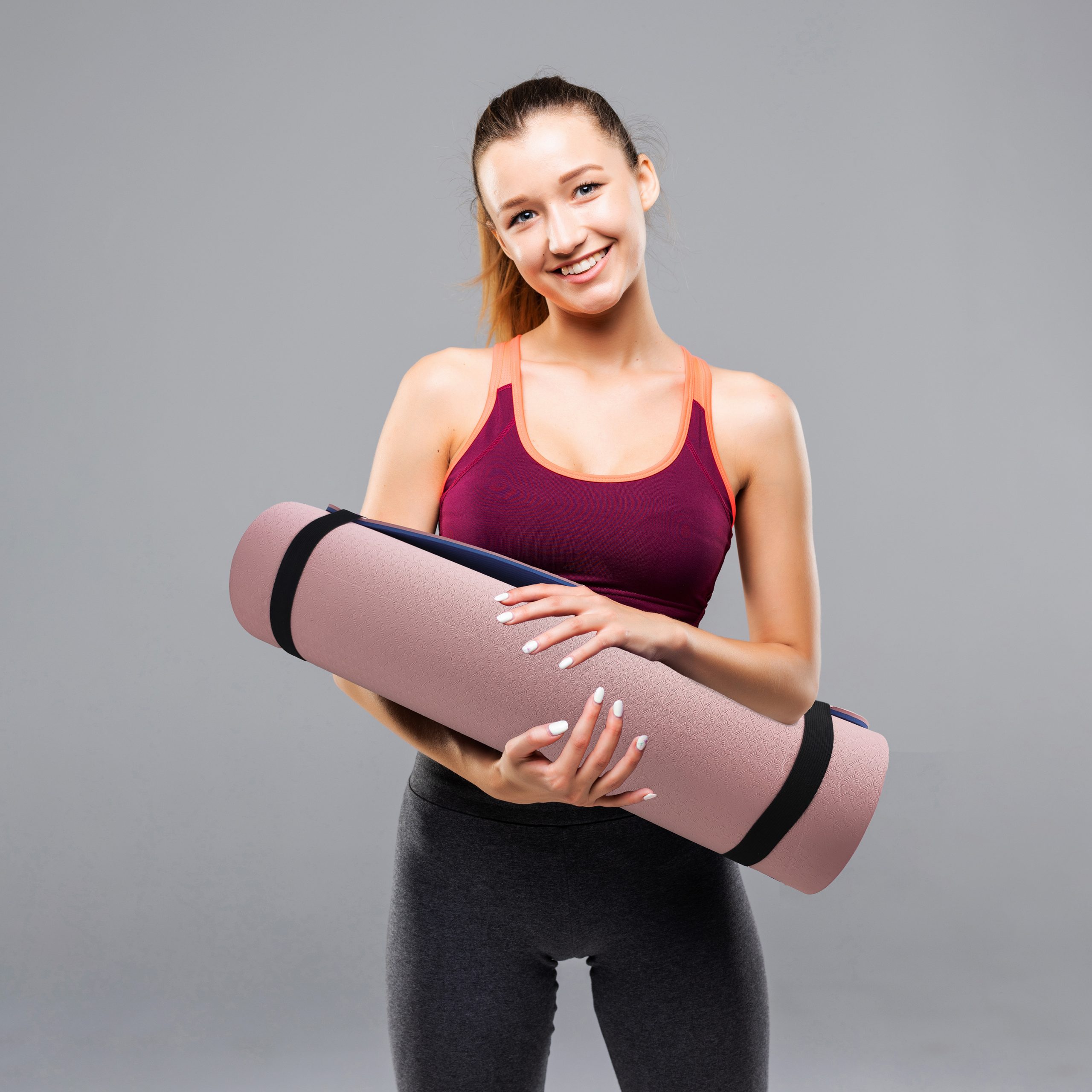 Smiling woman in athletic wear holding a rolled-up yoga mat against a gray background.