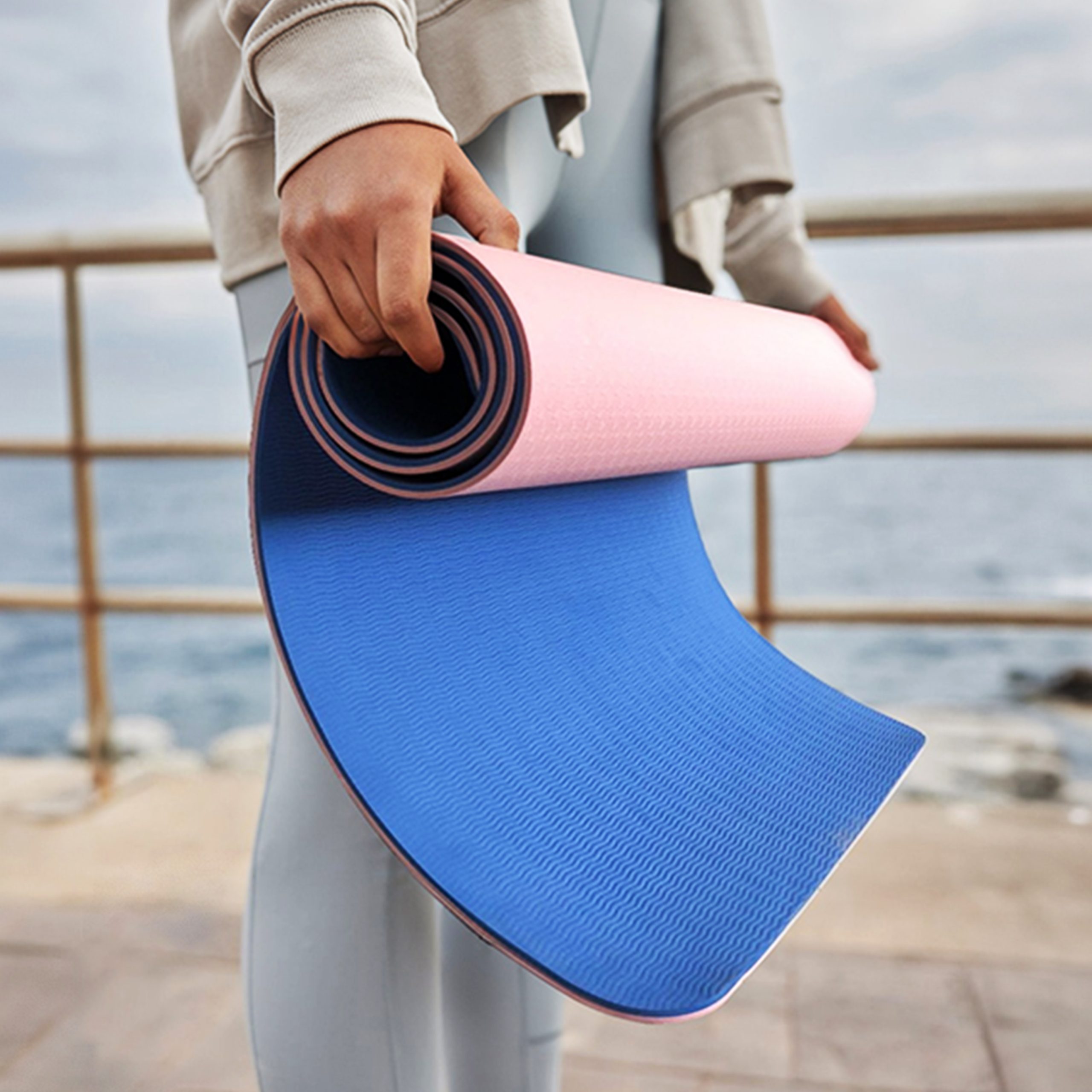 Hands unrolling a pink and blue yoga mat outdoors near water.