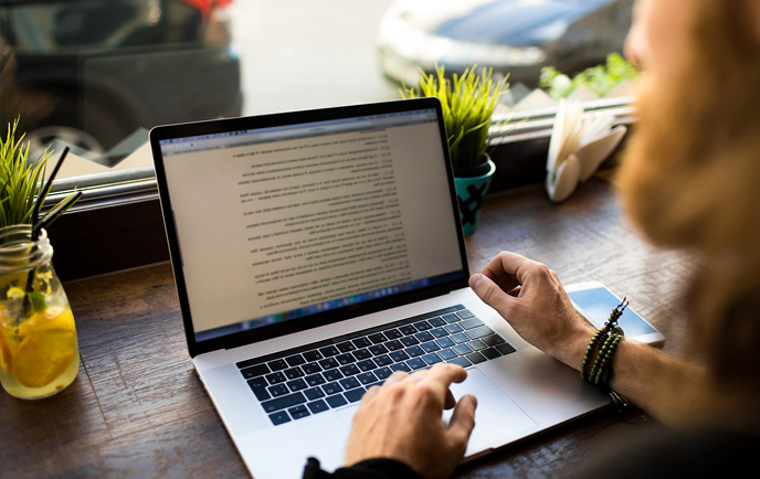 Person working on a laptop with text displayed on the screen, near a window and a drink.