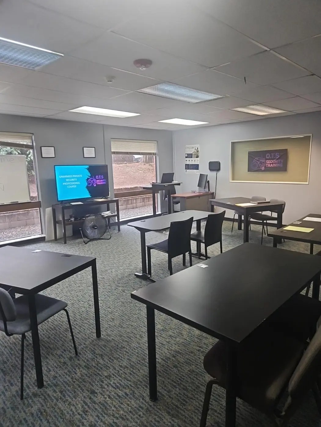 Wide view of the OTS Security Training classroom in Portland showing student tables, presentation screen, instructor workstation, and OTS wall display