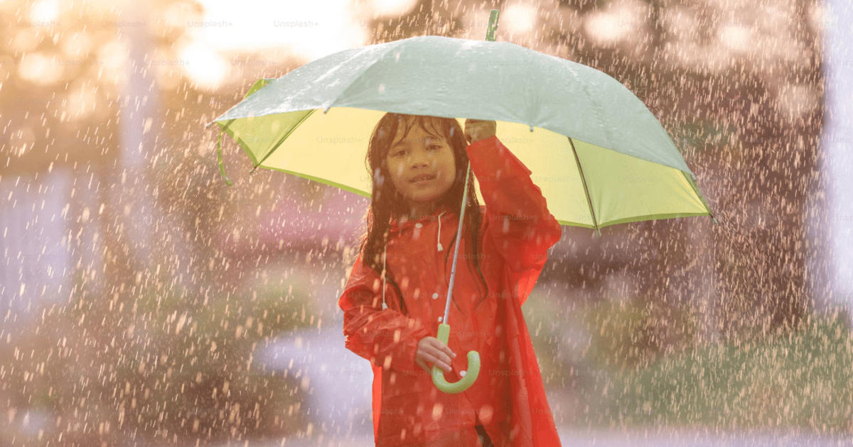 Little girl in orange raincoat with a tiny umbrella standing in the rain happily