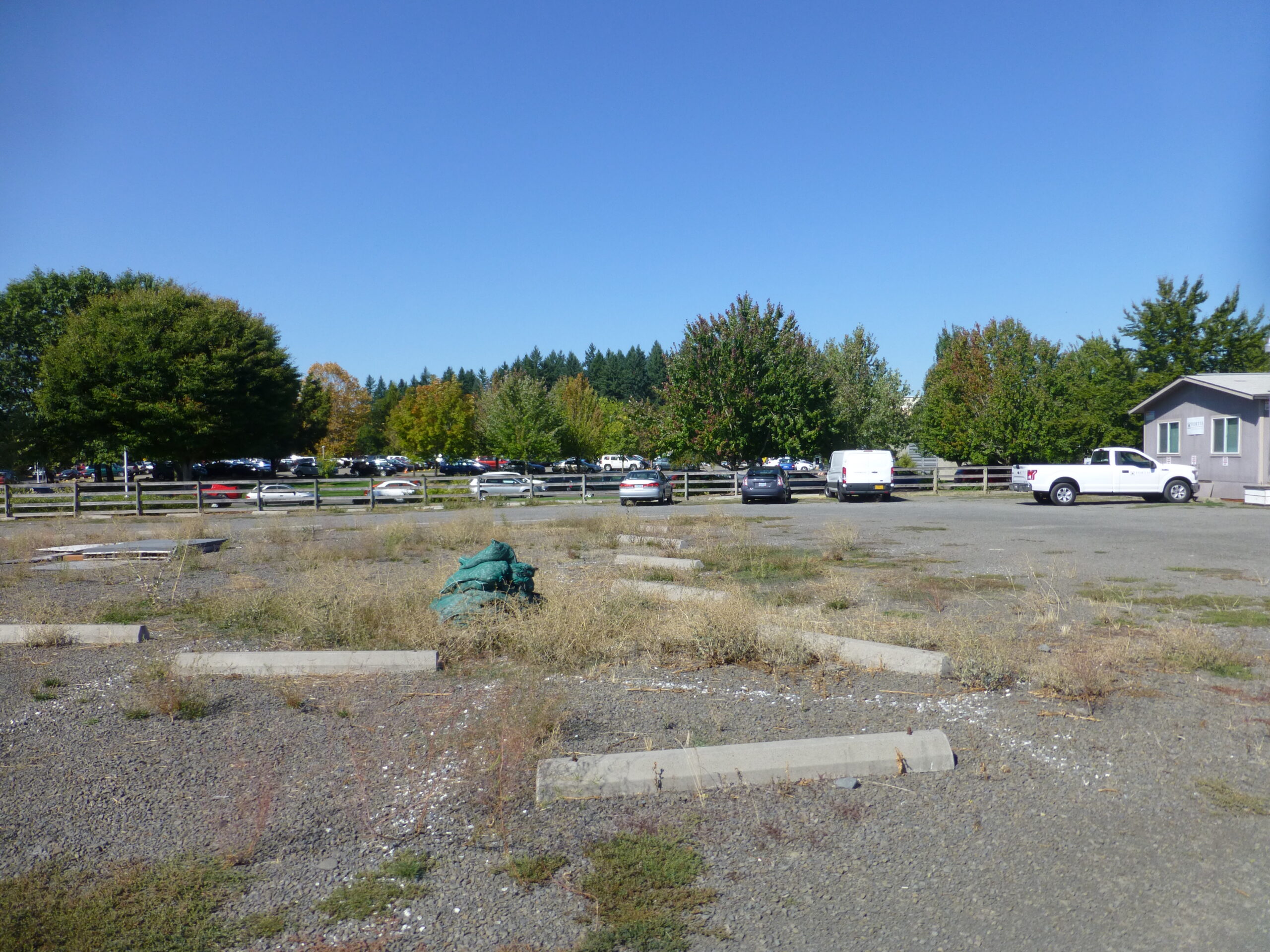 An empty gravel parking lot with overgrown weeds and scattered concrete parking stops, bordered by a wooden fence and rows of trees in the background. Several cars and trucks are parked near a small gray building on the right under a clear blue sky.
