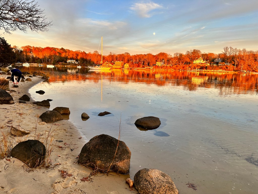 Cape Cod harbor at sunset with golden light, beach and stone foreground, and waterfront homes across the harbor representing Woods Hole and Upper Cape real estate