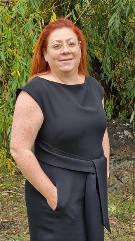 A woman with red hair and glasses stands in front of lush green trees, smiling softly at the camera