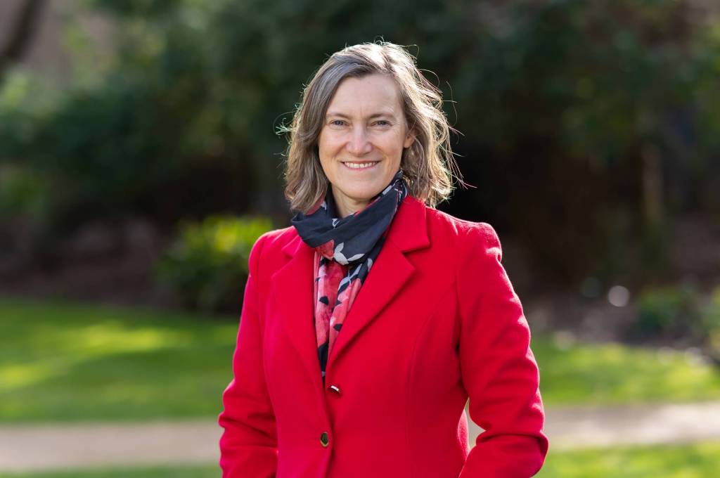 A woman in a red coat and scarf stands in a park, surrounded by trees and grass