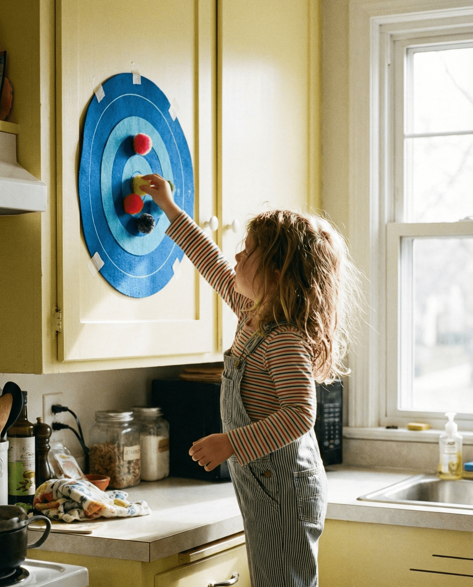 Preschool girl practices crossing midline by peeling pom-poms off a velcro felt target taped to a kitchen cabinet door for shoulder strengthening.