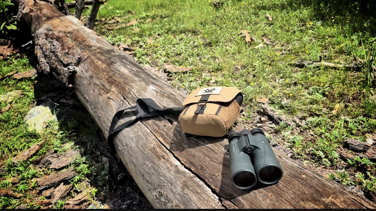 Full-size binoculars rest on a log with a chest harness.