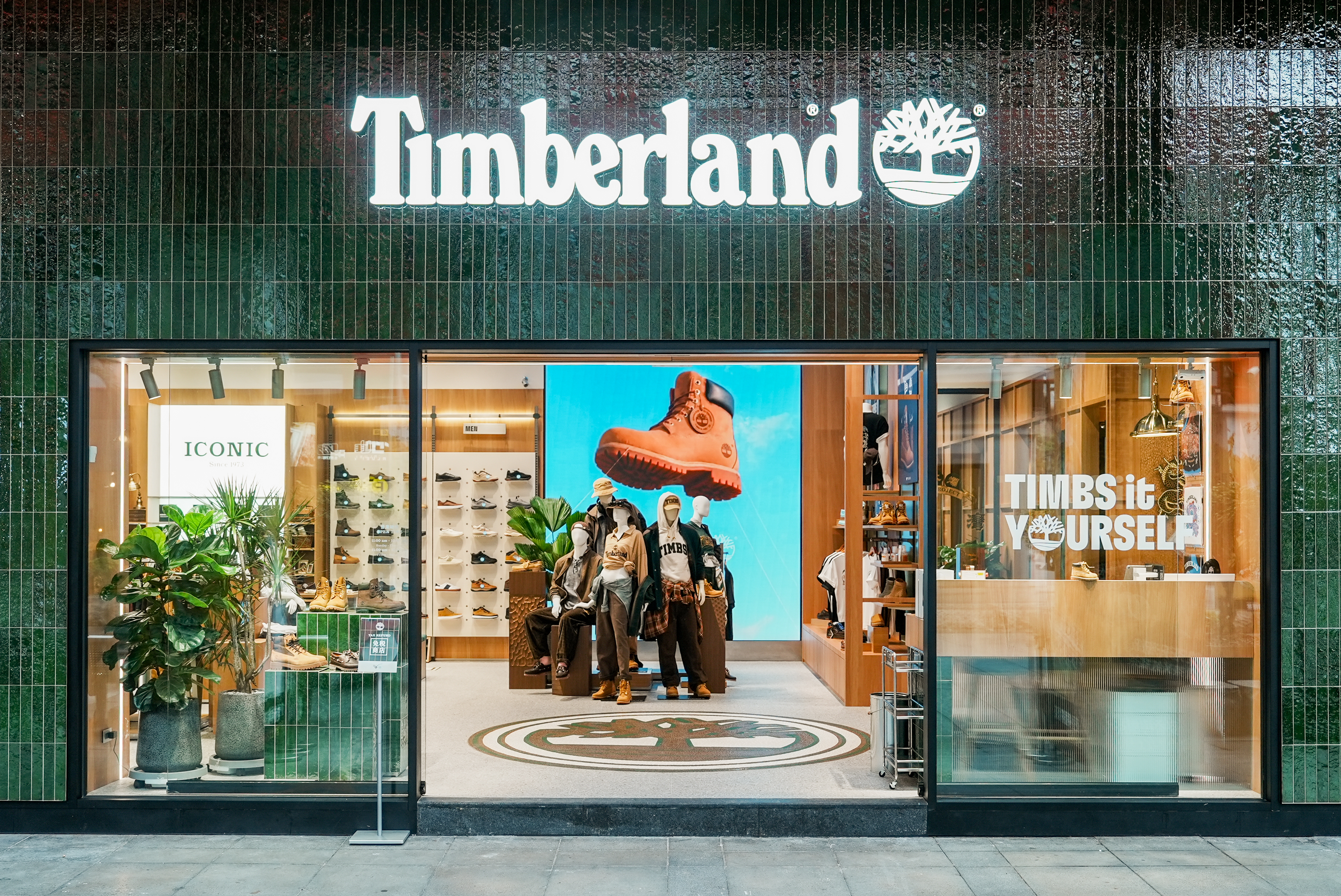 Timberland商店外觀，展示時尚的鞋子和服裝，橱窗内有模特和綠色植物，背景有大型藍色展示屏幕。