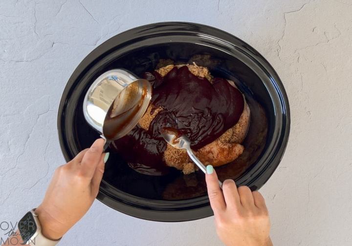 A black crock pot with seasoned chicken breasts layered in the bottom, topped with brown sugar and bbq sauce.