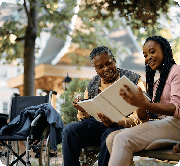 A smiling senior Black man and young Black woman share a book on a park bench.