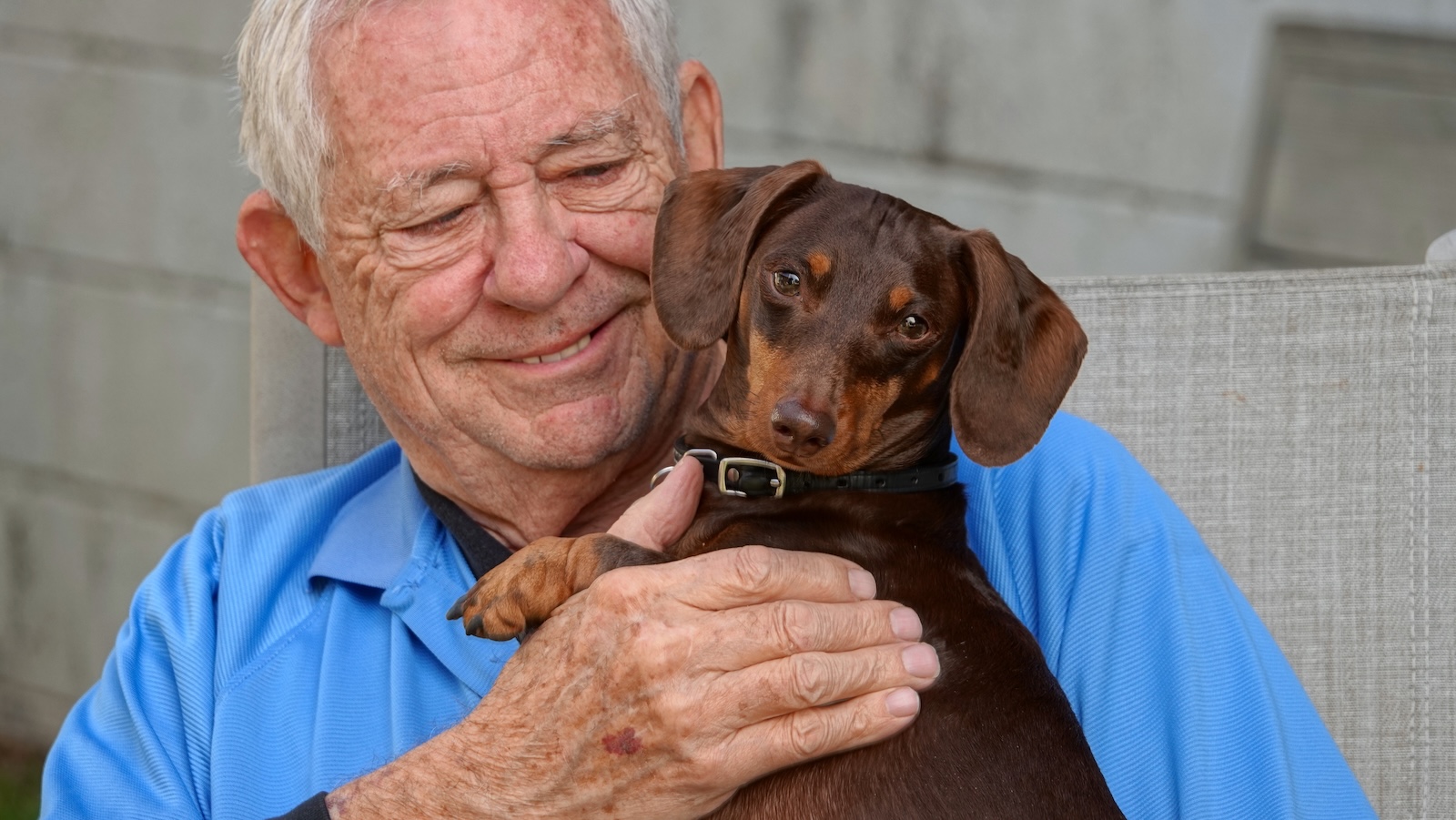 A happy senior man embraces his small brown dachshund with a gentle smile. TITLE: