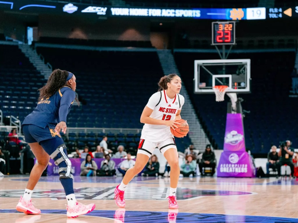A female basketball player from NC State dribbles the ball on the court while another player defends against her.