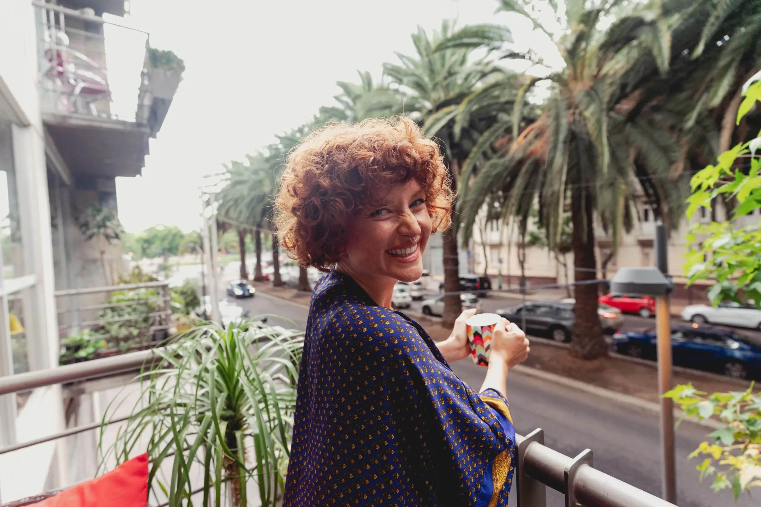 Woman with short curly hair looks at the camera smiling from a balcony outside.