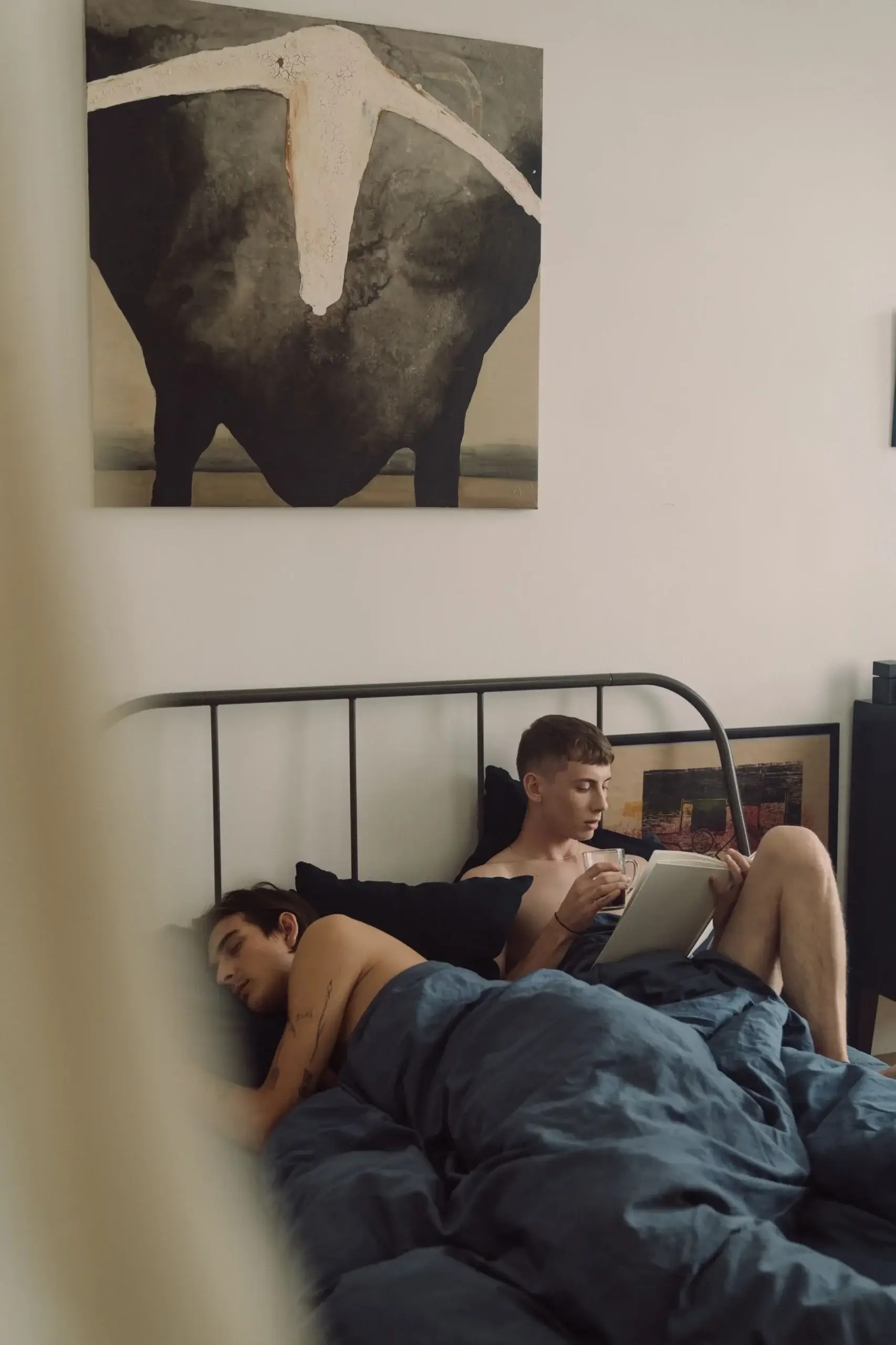 Two male partners lying in bed, with one sleeping and the other reading, illustrating different needs for closeness and space.